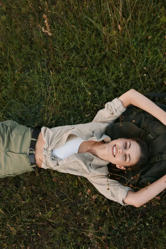 Relaxed woman lying on grass in casual outfit smiling, enjoying outdoor leisure under natural daylight, a warm lifestyle moment captured in a sunlit meadow.