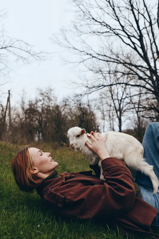 Joyful woman outdoors lying on green grass, smiling at a playful goat kid, warm sunlight, cozy jacket, relaxed interaction, natural colors, lush background, candid lifestyle moment