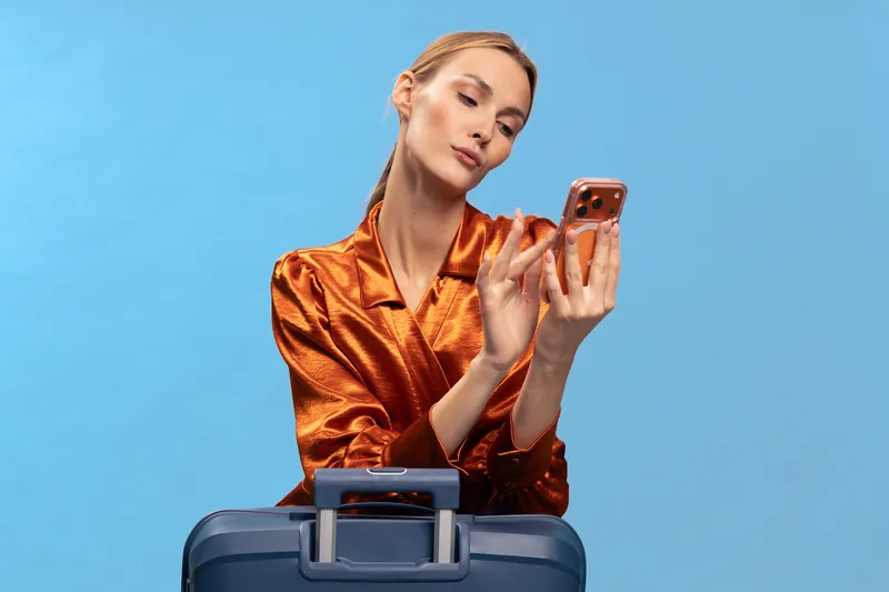 Young woman in shiny orange blouse checks smartphone while standing behind blue suitcase on blue background. Concept of travel planning, technology use, and modern lifestyle.