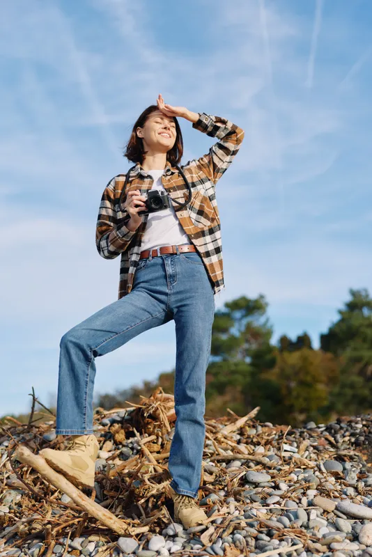 Woman with camera posing on rocky terrain under a clear blue sky, exploring nature and outdoor photography with a confident stance and casual outfit in bright daylight