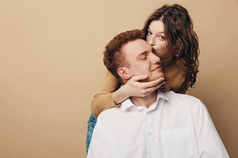 Young woman kisses man on cheek while holding his face gently. Couple shows affection and love in casual clothes on beige background. Intimate moment of tenderness and connection.