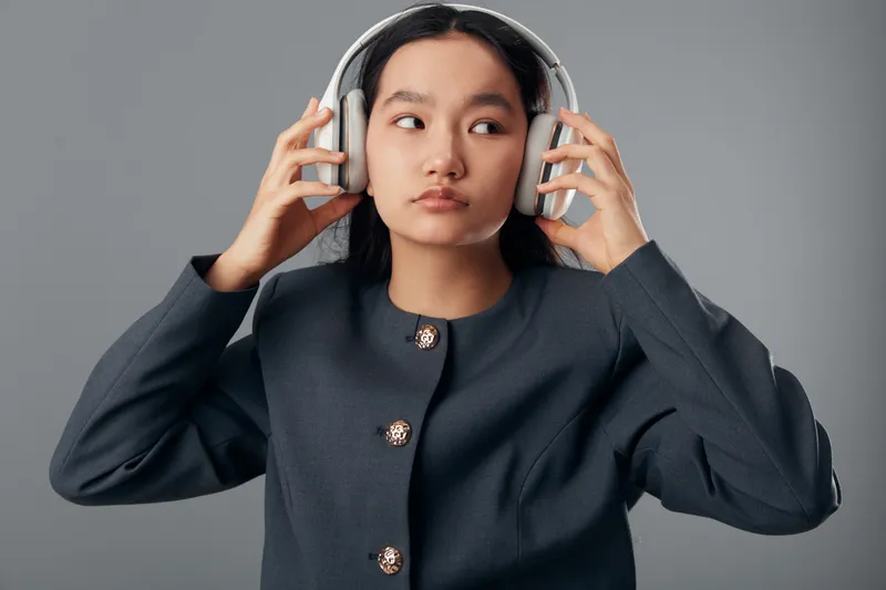 Young woman with headphones wearing dark jacket listens to music or audio with focused expression on grey background. Asian female adjusts accessory for sound experience indoors.