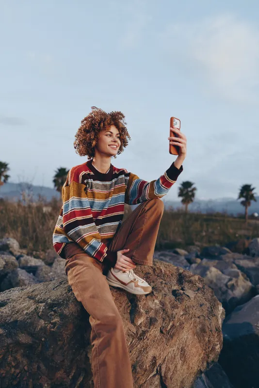 Woman lifestyle outdoors, sitting on a rock in a dry field, holding a phone for a selfie, wearing a casual striped sweater, curly hair in the wind, warm sunset light, scenic background
