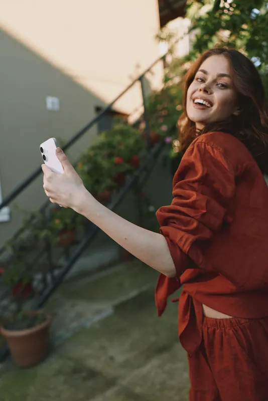 Cheerful woman outdoors, smiling while taking a selfie, casual rust outfit, sunlit moment, relaxed pose, soft breeze, natural colors, lush greenery in the background