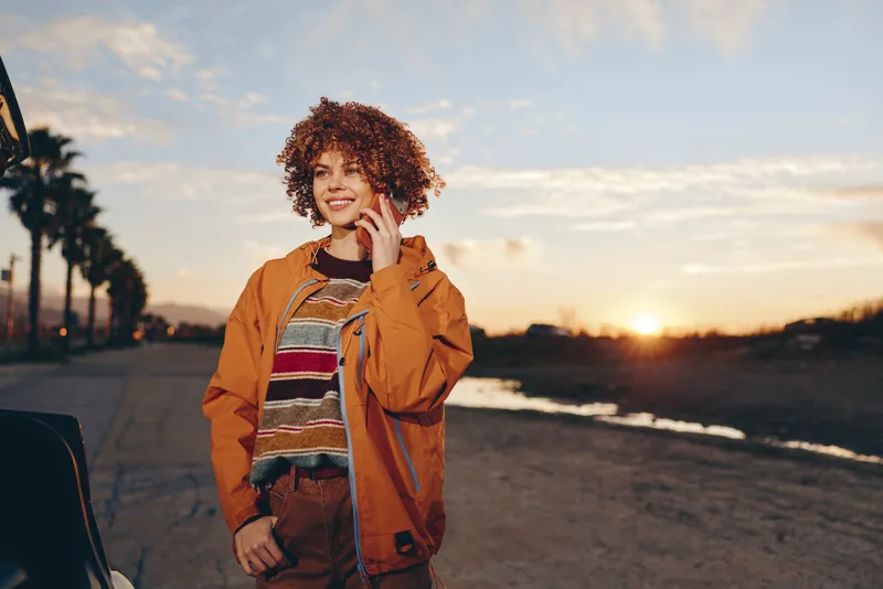 Curly haired woman lifestyle posing outdoors by the beach at sunset, wearing a warm jacket, smiling softly, golden light and ocean waves create a relaxed travel mood for everyday freedom