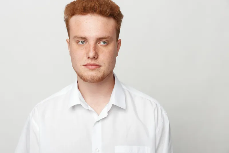 Young man with red hair and beard wearing white shirt looks away thoughtfully on light background. Concept of contemplation, casual style, and natural portrait.