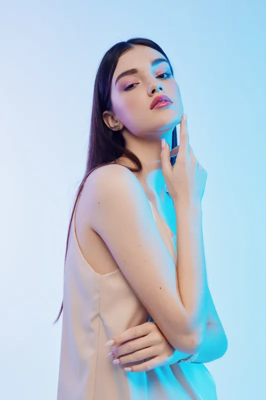 Young woman with long hair poses in a beige dress under soft blue light. Her hand gently touches her neck. Concept of woman beauty fashion and elegance in studio.