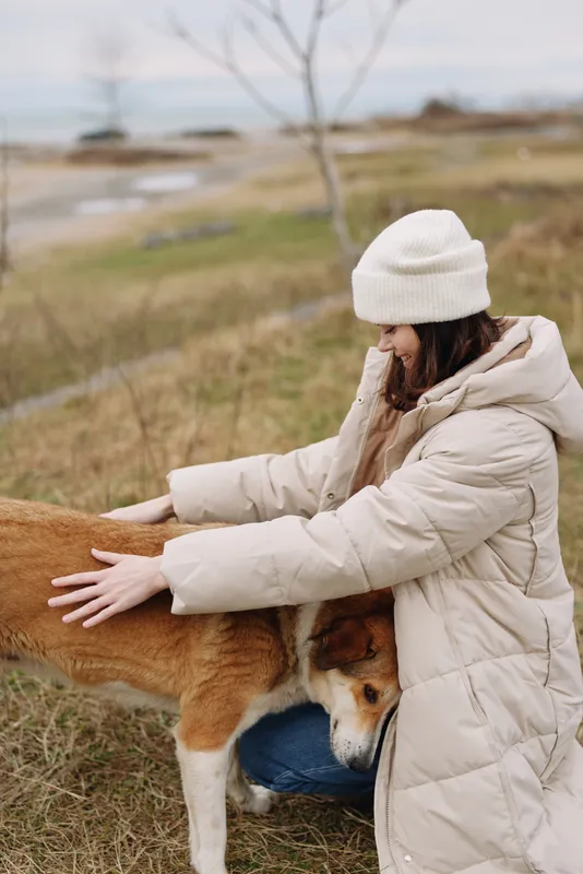 Dog, woman, outdoor, petting, winter, coat, field, companionship, a woman in a warm padded coat pats a calm dog in a vast grassy field under a pale sky, peaceful nature moment