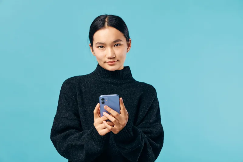 Woman holding smartphone with focus and calm expression on blue background. Female in black sweater using mobile phone for communication, technology and social media connection.