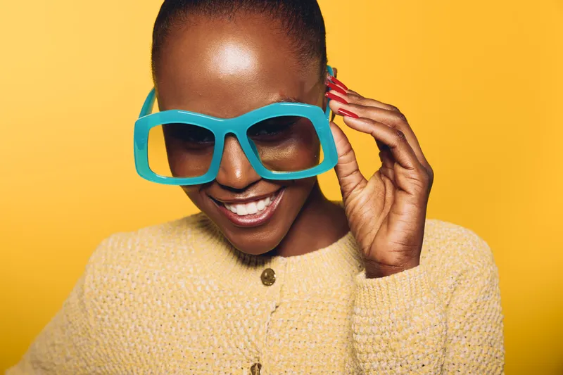 Smiling black African woman with dark skin wearing oversized blue glasses and yellow knitted sweater on yellow background. Concept of fashion, joy, style, and confident beauty.