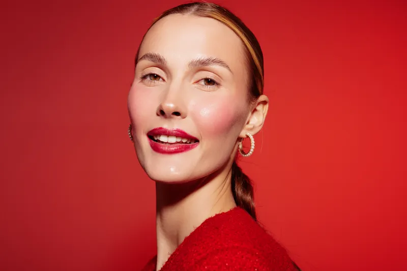 Smiling woman with red lipstick and hoop earrings poses against red background. Close-up portrait with natural makeup and sleek hairstyle. Concept of beauty, fashion, and confidence.