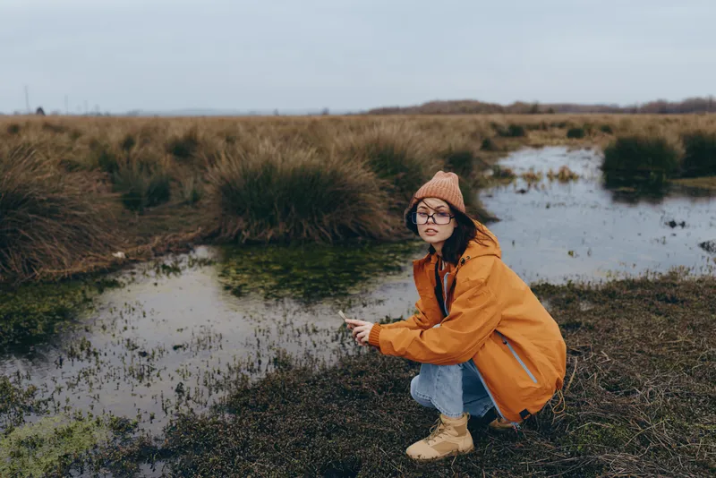 In autumn outdoors, a woman in an orange jacket crouches by a quiet river wetland marsh, surrounded by nature and tall grass, lifestyle moment with soft cloudy light in distance