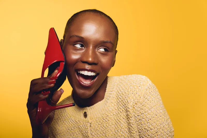 Happy African woman with dark skin holding red high heel shoe near face, smiling and looking aside. Bright yellow background highlights joyful mood and fashion concept with casual sweater.
