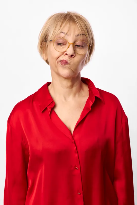Elderly woman with glasses, red shirt, isolated portrait on white studio background, pouting lips and squinting expression, displeased emotion