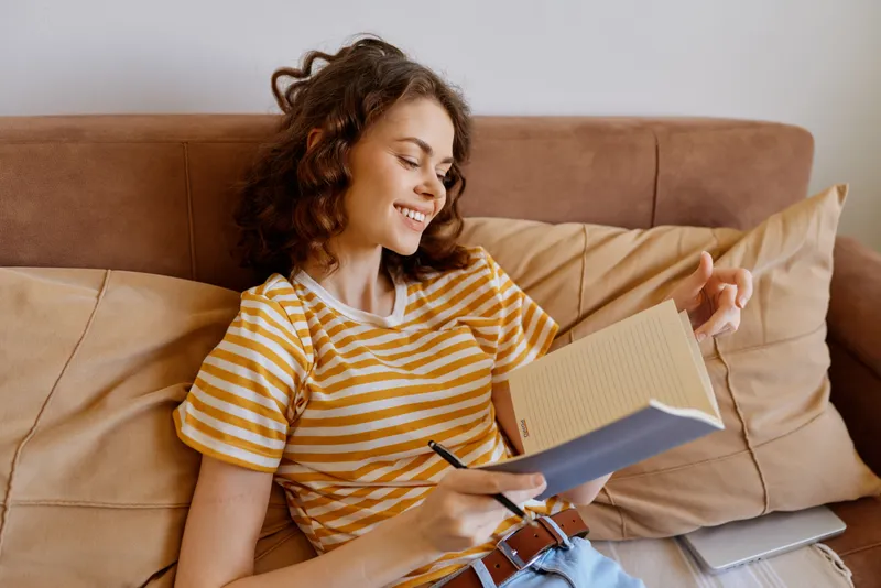 Young woman relaxing on a sofa at home, writing in a notebook and smiling as she sketches plans for the week.