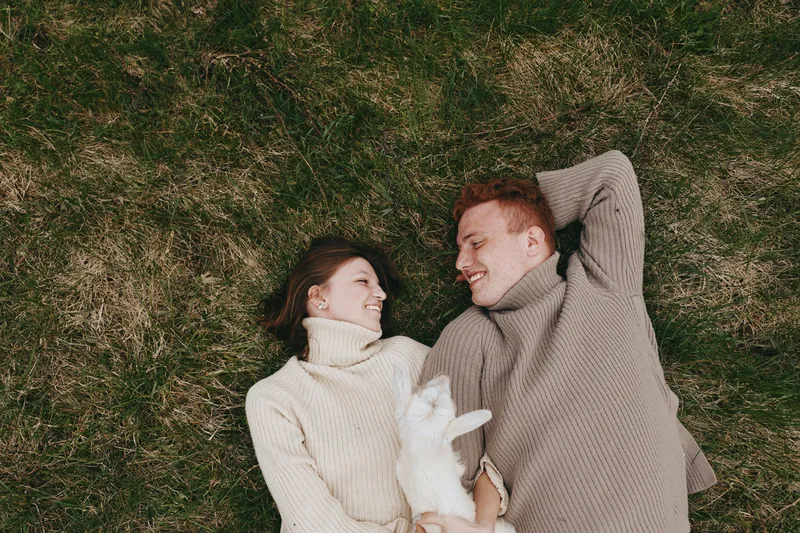 Young couple in cozy sweaters lies on grass smiling at each other while holding a small white baby goat in a relaxed outdoor lifestyle moment.