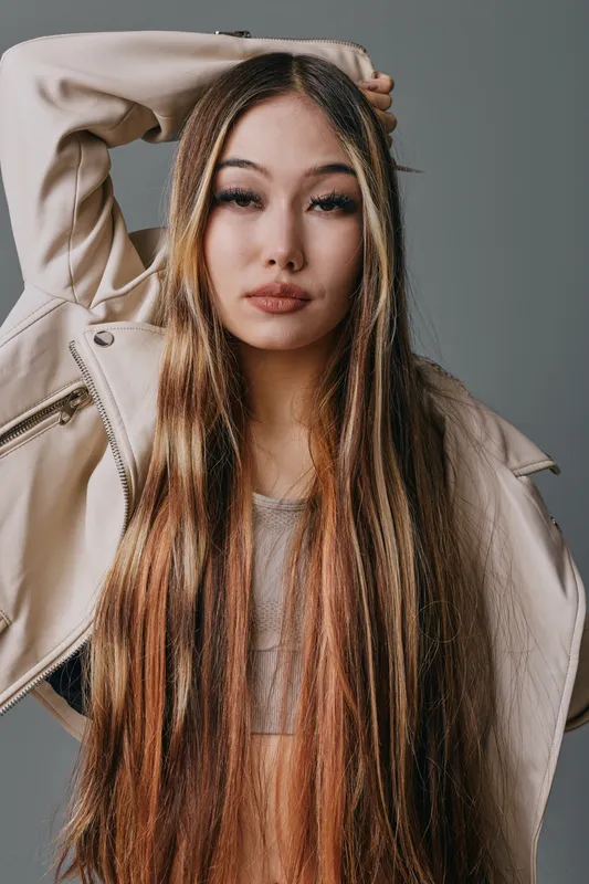 Young woman with long straight hair wearing beige jacket posing with one arm raised on neutral background. Fashion portrait, confident expression, casual style for beauty and lifestyle.