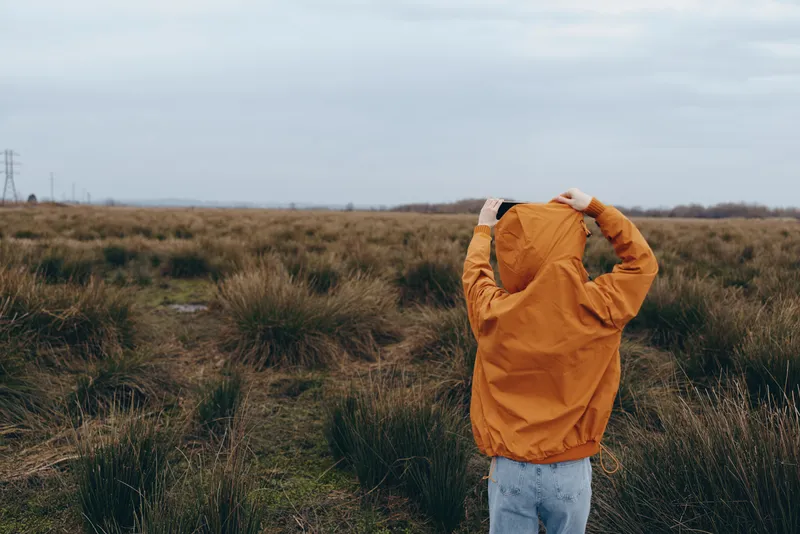 Woman lifestyle outdoors meadow under cloudy sky, standing in tall grass field, holding orange jacket overhead, relaxed travel mood, quiet morning wind, wide nature landscape background