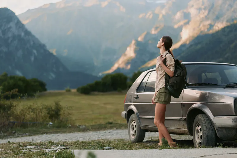 Woman traveler with backpack stands beside a car in a rugged mountain landscape, enjoying fresh air and open scenery, ready for a hiking adventure