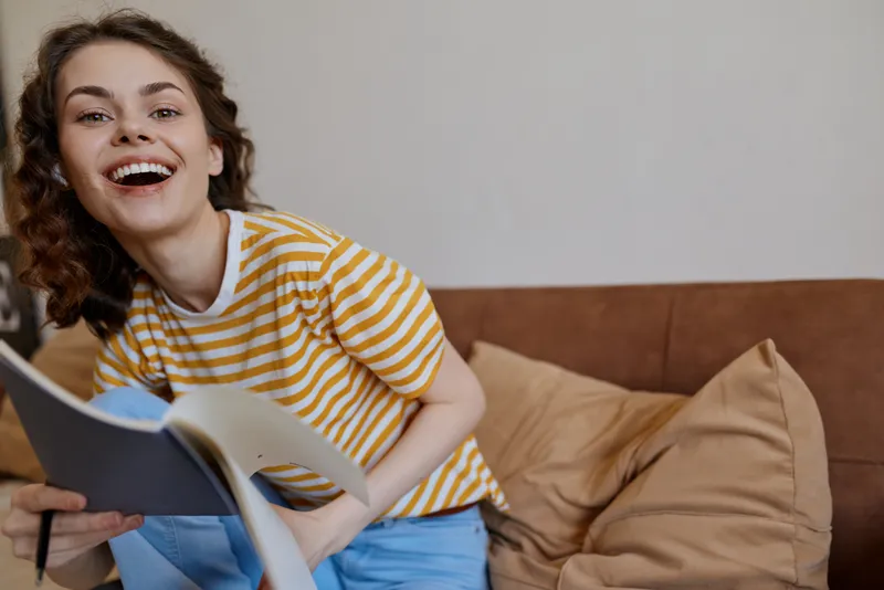 A joyful woman sits on a sofa at home, reading a book with a wide smile. A warm, casual lifestyle moment focused on comfort, everyday reading, and self care.