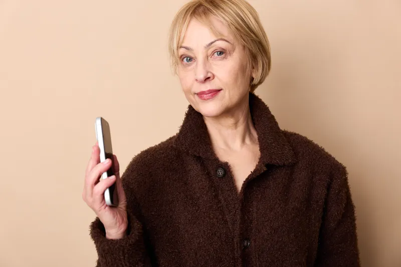 Woman holds a smartphone in a studio portrait of a senior female with calm smile and warm coat, looking at camera, concept of technology communication and indoor lifestyle indoors
