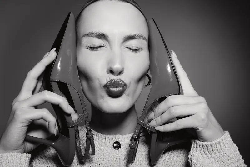 Caucasian woman with light skin holds pair of high heel shoes near face and makes kiss gesture. Studio portrait in black and white with fashion and beauty concept.