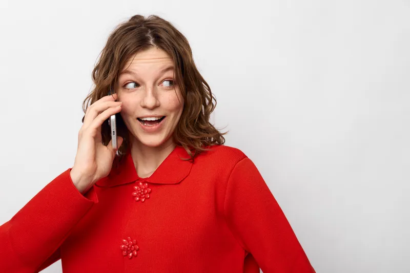 Young woman in red sweater talks on smartphone with surprised expression, looking aside. Isolated on white background. Concept of communication, technology, and casual lifestyle.