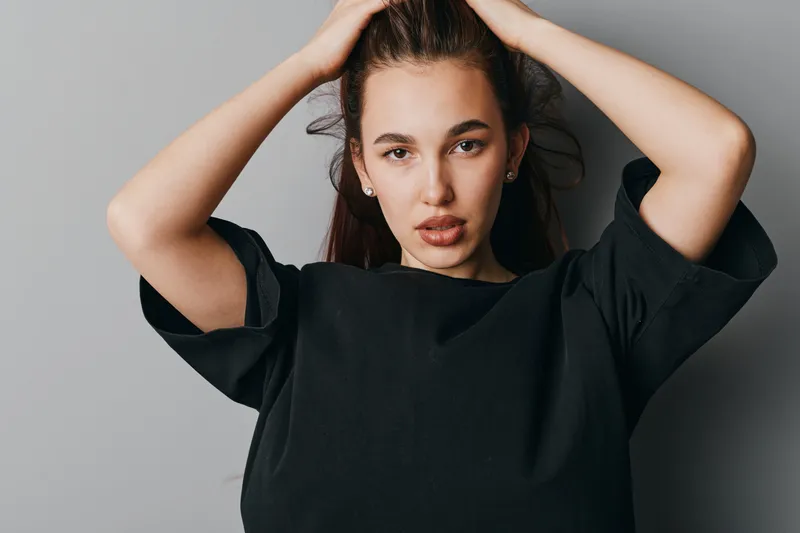 Young woman with long dark hair wearing black shirt posing with hands on head against gray background. Portrait of confident female model with natural makeup and serious expression.