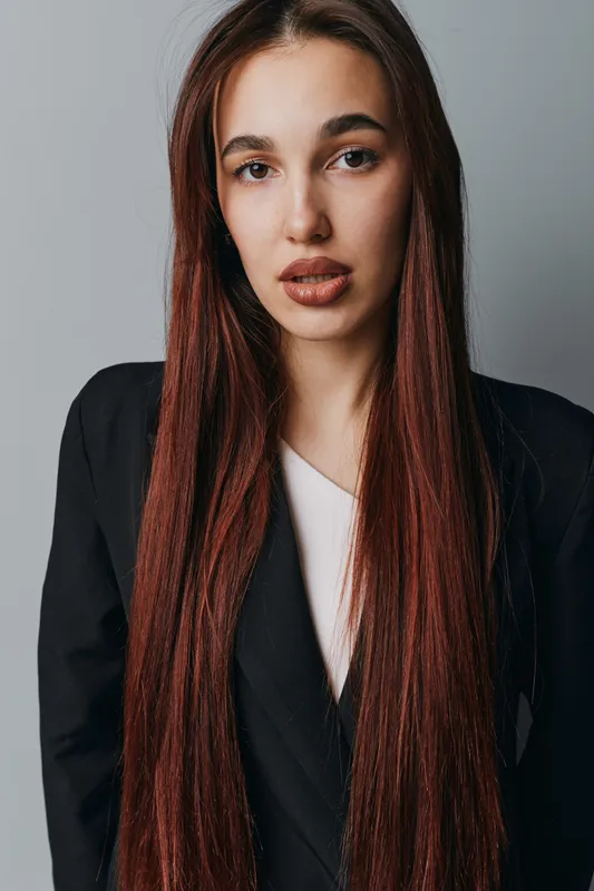 Young woman with long straight hair wearing black blazer and white shirt posing against gray background. Portrait of confident female model with natural makeup and serious expression.