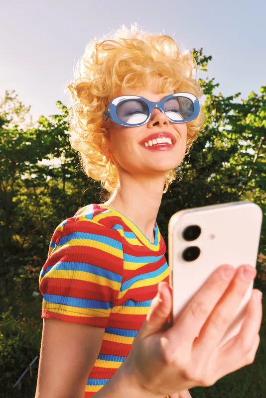 Retro selfie of a smiling woman with curly blond hair, striped shirt, and sunglasses, outdoors in bright sunny weather. Portrait mood, cheerful fashion vibe, color mood, vintage flair, and carefree