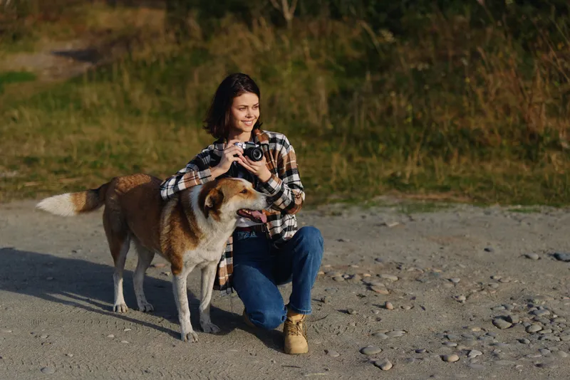 Woman kneels beside a dog in a rocky outdoor scene, using a camera to capture moments in nature with casual clothing and warm daylight, creating a candid travel portrait.