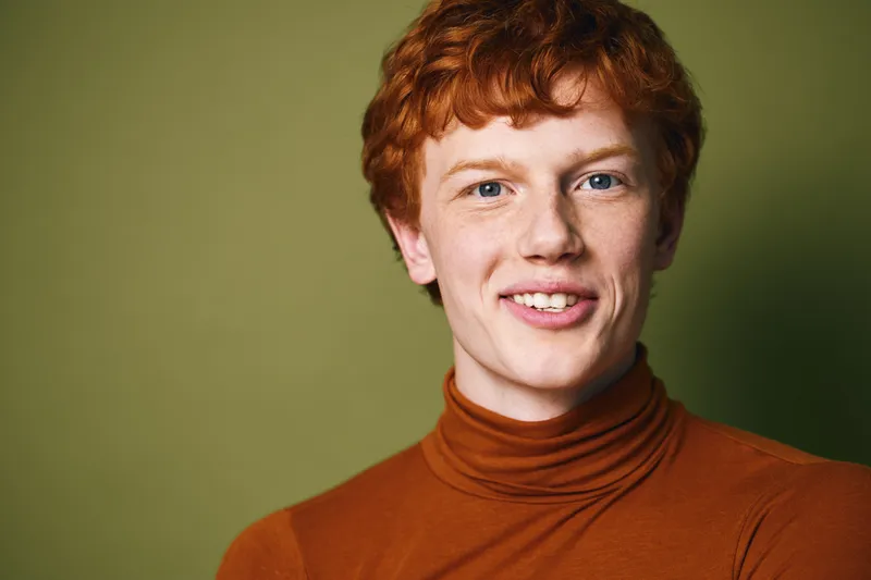 Young man with red hair and freckles smiling wearing orange turtleneck sweater on green background. Closeup portrait with natural expression and casual style in studio lighting.