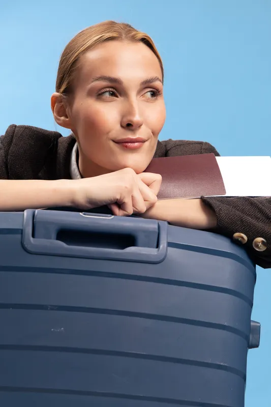 Young woman leans on blue suitcase holding a brown notebook, looking sideways with slight smile. Travel concept with relaxed attitude and modern casual style on light blue background.