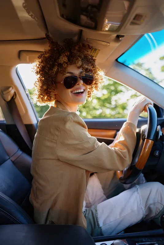 Smiling woman with curly hair driving a car, wearing sunglasses, enjoying a sunny day. Casual fashion, relaxed lifestyle, and a candid moment inside a warm vehicle.
