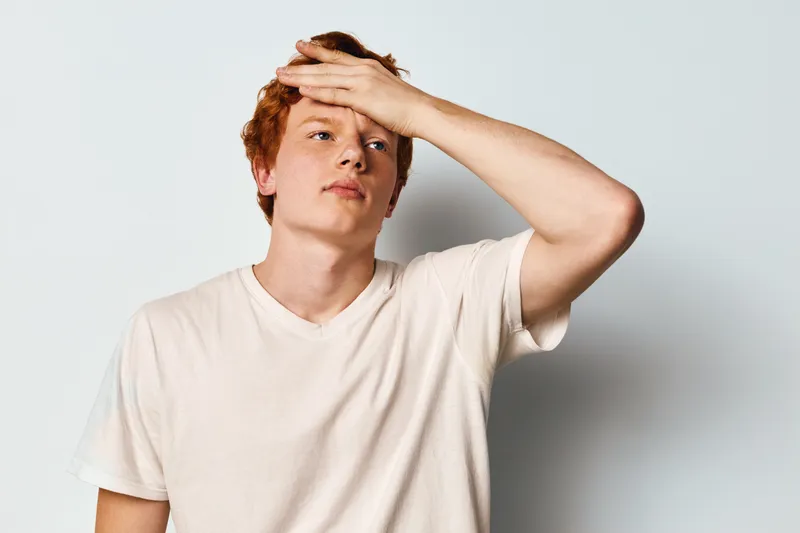 Young man with red hair in white t shirt touching forehead and looking away with tired expression against light background. Casual portrait of male showing fatigue or stress.