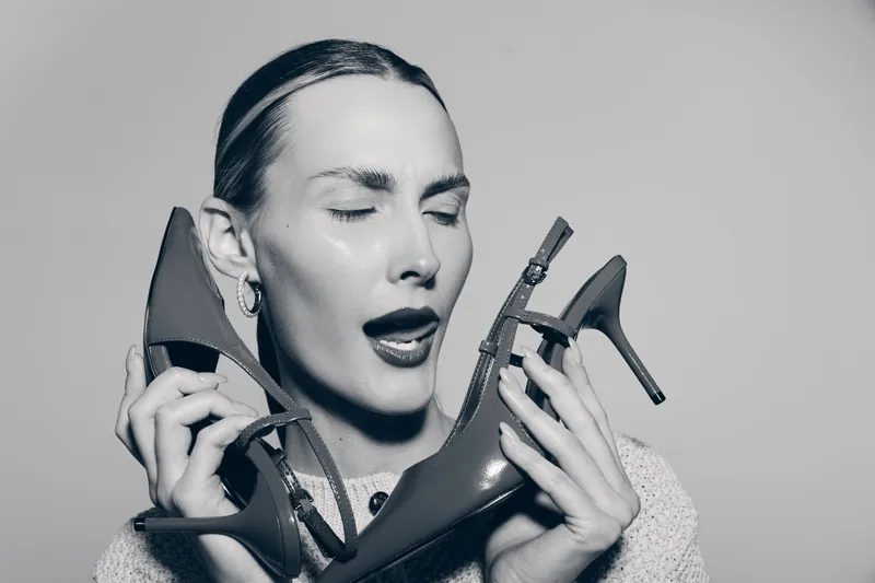 Young Caucasian woman with fair skin holds two high heel shoes near her face with closed eyes and playful expression. Concept for fashion, footwear, and personal style.