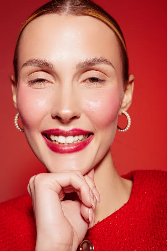 Close-up portrait of smiling woman with red lipstick and hoop earrings against red background. Concept of beauty, makeup, fashion, and confident female model with natural skin glow.