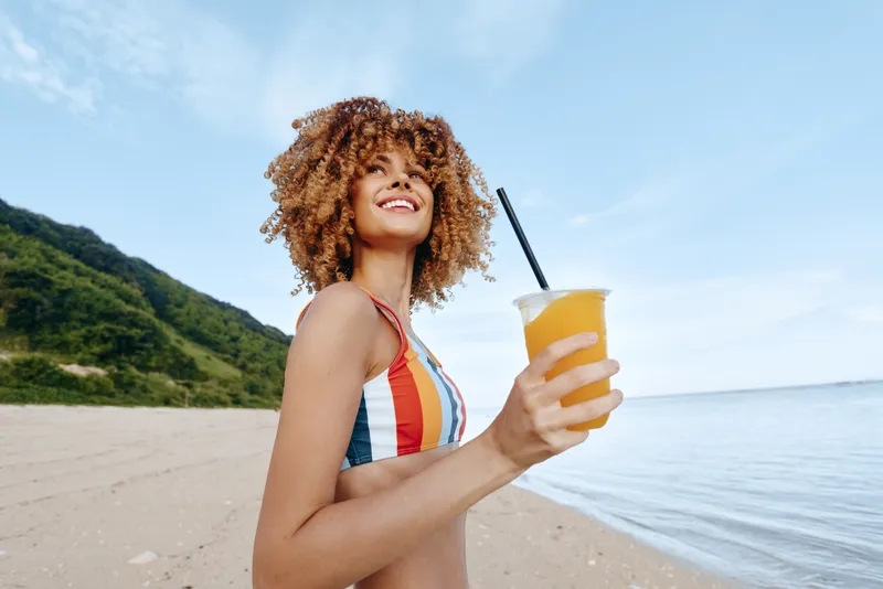 Woman on beach by ocean holding juice in glass smiles in summer, curly hair blowing, relaxed seaside mood, holding drink, sunny vacation light, natural outdoor background, copy space