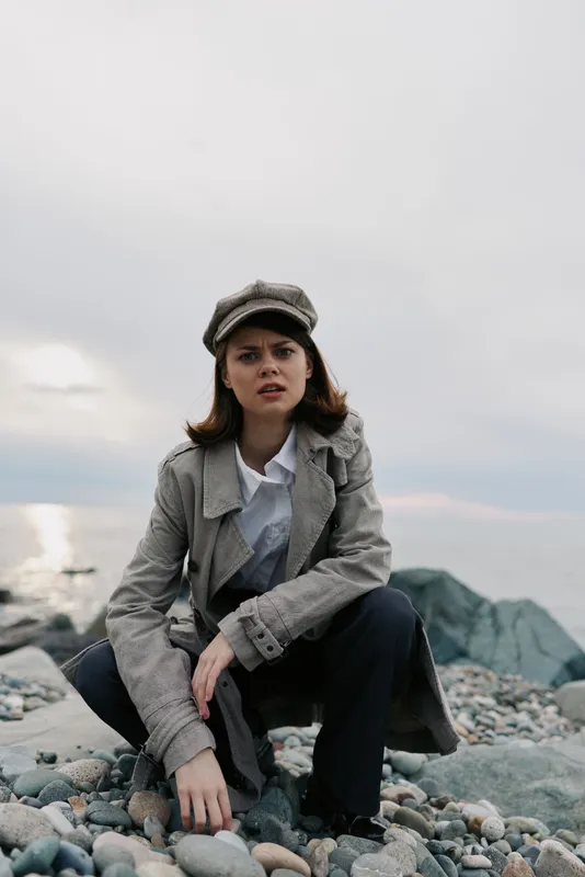 Young woman in vintage coat and cap crouches on rocky beach with cloudy sky and calm sea behind. Concept for fashion, autumn style, and outdoor portrait photography.