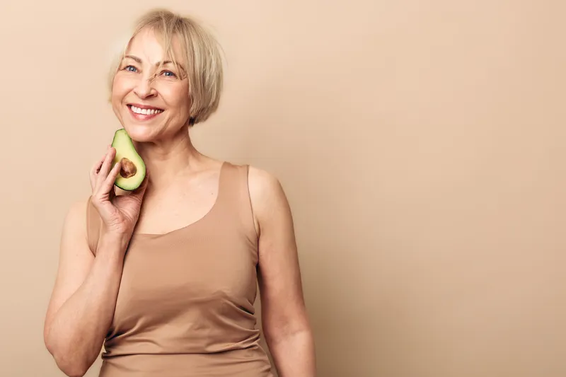 Smiling mature woman holds half avocado near face, wearing beige tank top against neutral background. Concept for healthy lifestyle, nutrition, and natural wellness.