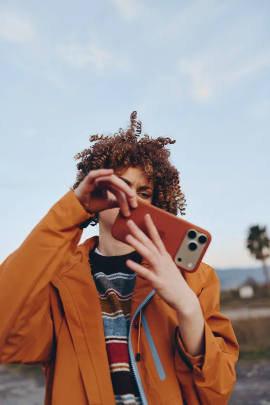 Woman lifestyle portrait outdoors, wearing orange jacket and curly hair, holding smartphone in hand, adjusting face with fingers, sunny sky background, calm confident mood today outside