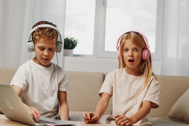 Curious boy and surprised girl with headphones use laptop on couch in cozy bright room. Concept for children learning, technology, and digital discovery.