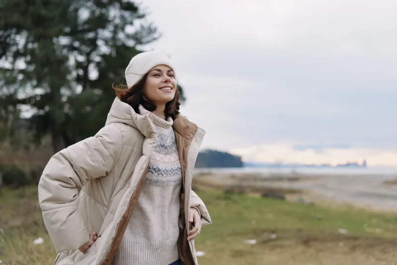 Woman outdoors wearing a cozy coat in a scenic beachside park, smiling and confident, knit sweater and beanie, breathing fresh air in natural light and calm surroundings