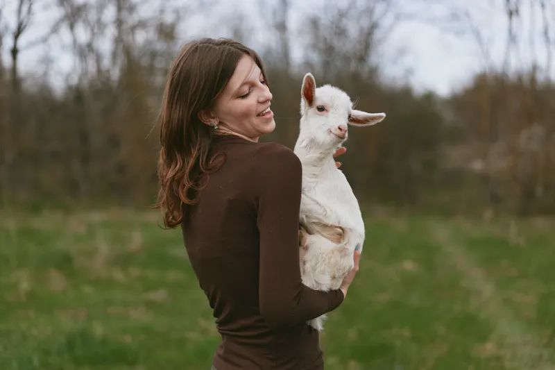 Young woman with long brown hair holds a white baby goat outdoors in a natural green field during a calm overcast day, showing affection and joy in a lifestyle moment.