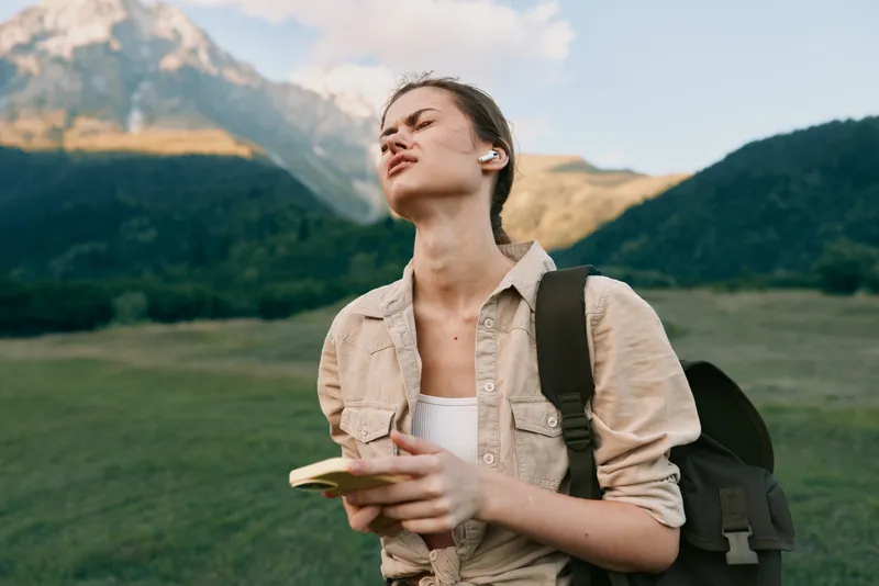 Young woman traveler in a green valley, notebook in hand, backpack over her shoulder, gazing toward distant mountains. Natural light highlights outdoor exploration and curiosity.