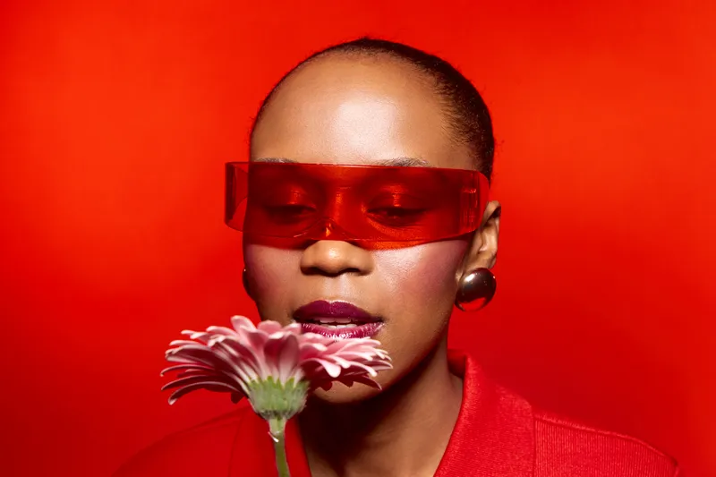 Portrait of dark skinned African woman wearing red sunglasses and red clothes holding pink flower on vibrant red background. Concept of fashion, beauty, and bold color style.