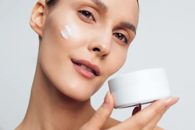 Female portrait applying moisturizer, close up of face with cream on cheek, holding jar, smooth skin, calm expression, minimal studio lighting, skincare routine concept, beauty care