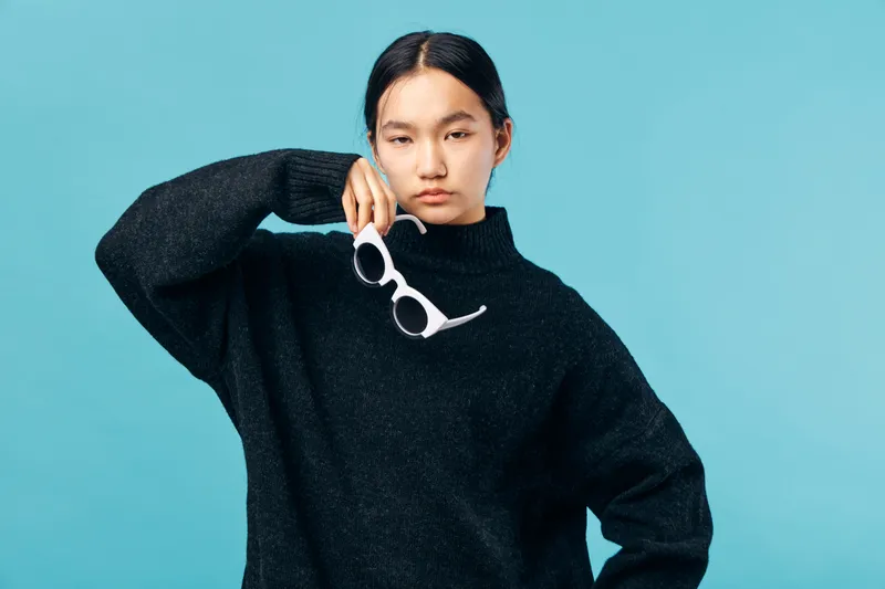 Woman posing with white sunglasses in hand wearing black sweater with high neck against blue background. Asian female model with serious expression and casual fashion style.