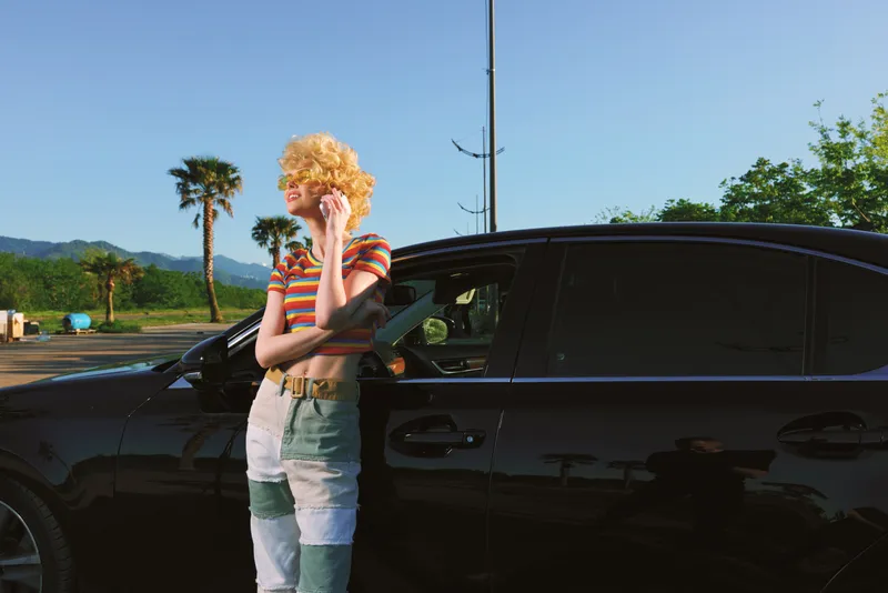 Cheerful woman outdoors beside a sleek car, striped shirt and denim jeans, sunlit moment with gentle breeze, relaxed posture, retro film color and natural mood with warm tones today