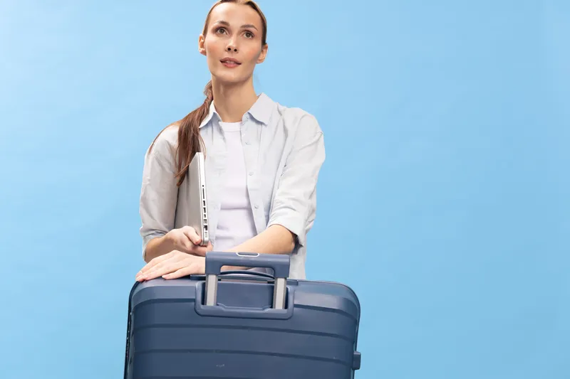 Young woman with long hair holding laptop and standing behind large suitcase on blue background. Concept of travel preparation, business trip, and modern lifestyle with technology.